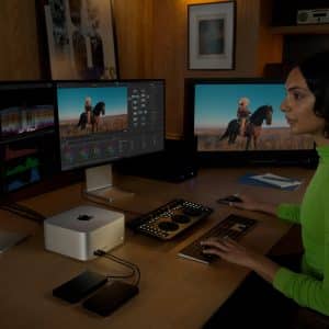 A person in a neon green shirt sits at a desk with a Mac Studio, editing video and color on a multi-monitor setup. The workspace features external drives, control panels, photos on the wall, and a shelf filled with books.
