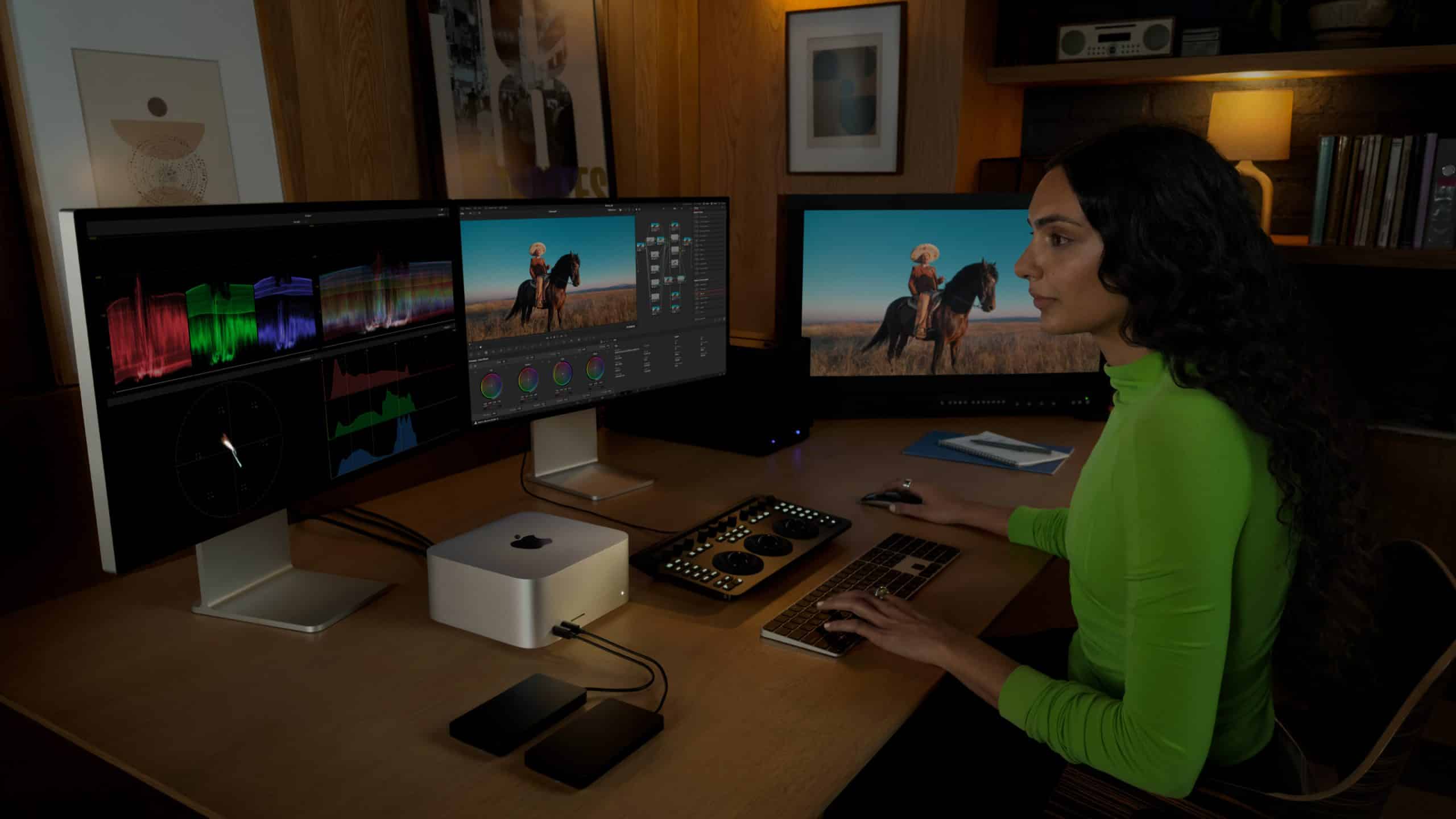 A person in a neon green shirt sits at a desk with a Mac Studio, editing video and color on a multi-monitor setup. The workspace features external drives, control panels, photos on the wall, and a shelf filled with books.