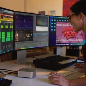 A person using a MacBook with three monitors displaying music production software. A MIDI keyboard and audio interface sit on the desk. Another person stands nearby, partially visible, while a poster about music production classes hangs in the background.
