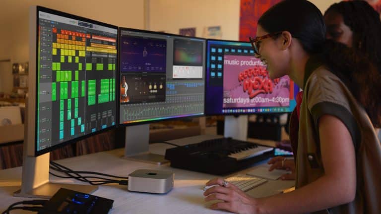 A person using a MacBook with three monitors displaying music production software. A MIDI keyboard and audio interface sit on the desk. Another person stands nearby, partially visible, while a poster about music production classes hangs in the background.
