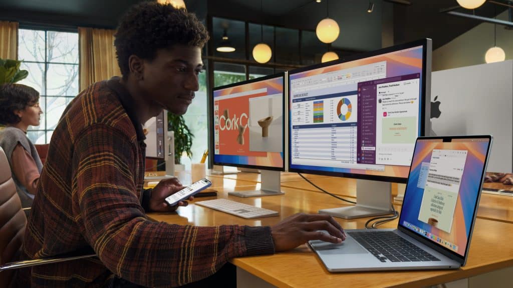 A person in a colorful sweater uses a laptop at a desk, contemplating severance details on their smartphone. They glance at one of two large monitors displaying work-related content. Another person works in the background, and the room has modern decor with round ceiling lights.