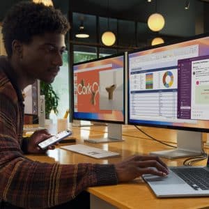 A person in a colorful sweater uses a laptop at a desk, contemplating severance details on their smartphone. They glance at one of two large monitors displaying work-related content. Another person works in the background, and the room has modern decor with round ceiling lights.