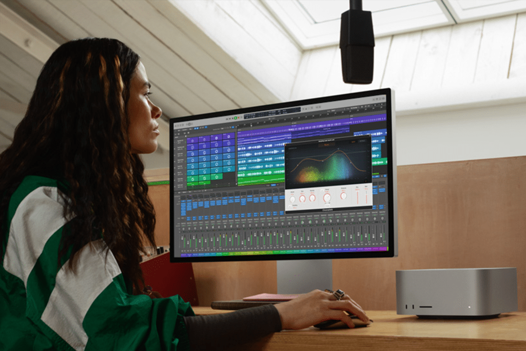 A person wearing a green and white jacket sits at a desk using an M4 Max computer with audio editing software displayed on the monitor. The room is well-lit with natural light streaming from a skylight above.