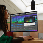 A person wearing a green and white jacket sits at a desk using an M4 Max computer with audio editing software displayed on the monitor. The room is well-lit with natural light streaming from a skylight above.