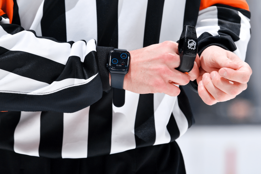 An NHL referee adjusts a strap on their left wrist, wearing the iconic black and white striped shirt. Their Apple Watch, a piece of cutting-edge technology, displays a digital face as the game buzzes in the blurred background.