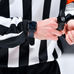 An NHL referee adjusts a strap on their left wrist, wearing the iconic black and white striped shirt. Their Apple Watch, a piece of cutting-edge technology, displays a digital face as the game buzzes in the blurred background.