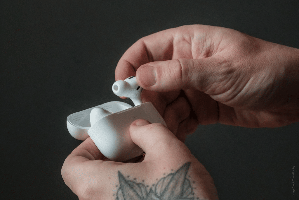 A person holding white AirPods with one hand carefully places them into a charging case with the other. A tattoo of a butterfly graces their wrist, adding a touch of elegance to the scene against the plain, dark background.