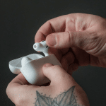 A person holding white AirPods with one hand carefully places them into a charging case with the other. A tattoo of a butterfly graces their wrist, adding a touch of elegance to the scene against the plain, dark background.