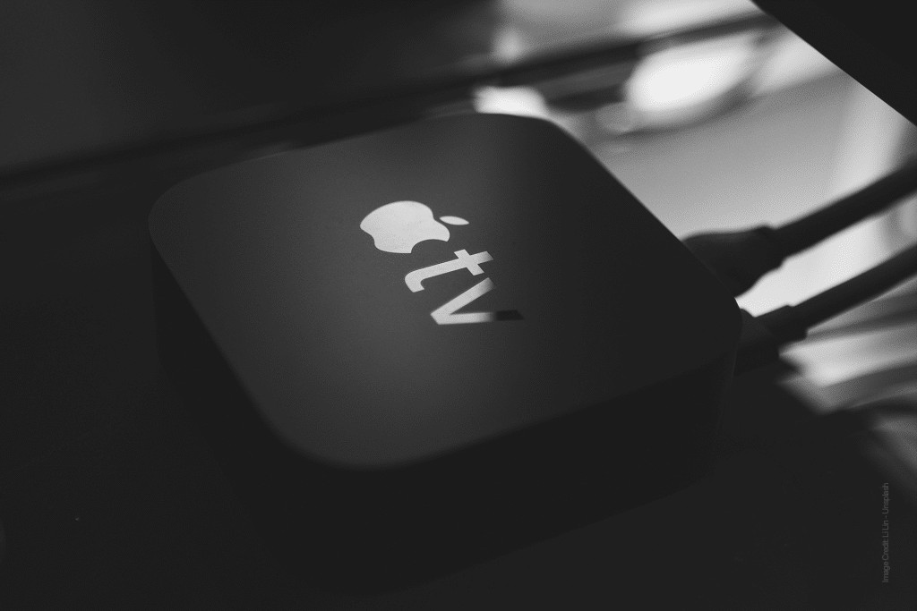 A close-up black and white image of the Apple TV 4K device on a surface. The sleek gadget features the iconic Apple logo with "tv" beneath it, and its advanced 4K capabilities ensure an immersive experience. Cables snake discreetly from its back, ready for entertainment.