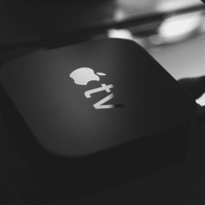 A close-up black and white image of the Apple TV 4K device on a surface. The sleek gadget features the iconic Apple logo with "tv" beneath it, and its advanced 4K capabilities ensure an immersive experience. Cables snake discreetly from its back, ready for entertainment.