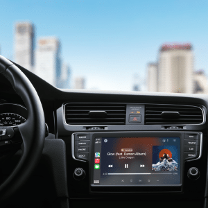 View from a car's interior featuring a CarPlay digital display playing "Glow" by Little Dragon. The city skyline with towering buildings stretches through the windshield, set against a clear blue sky.