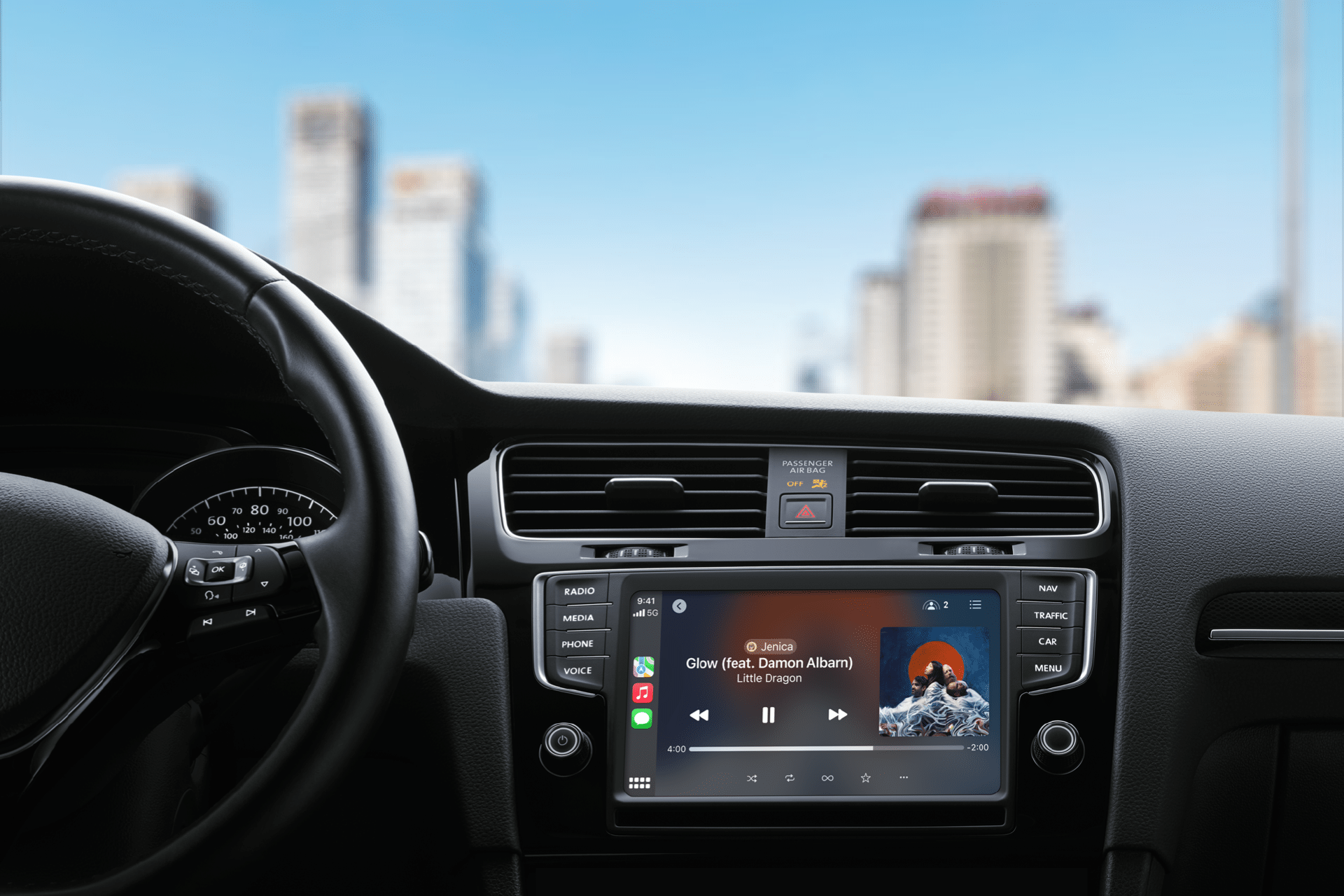 View from a car's interior featuring a CarPlay digital display playing "Glow" by Little Dragon. The city skyline with towering buildings stretches through the windshield, set against a clear blue sky.