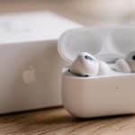 A pair of sleek, white wireless earbuds rest elegantly in their open charging case on a wooden surface, ready to fill the air with sound. In the background, a blurred box with an Apple logo hints at refined quality.