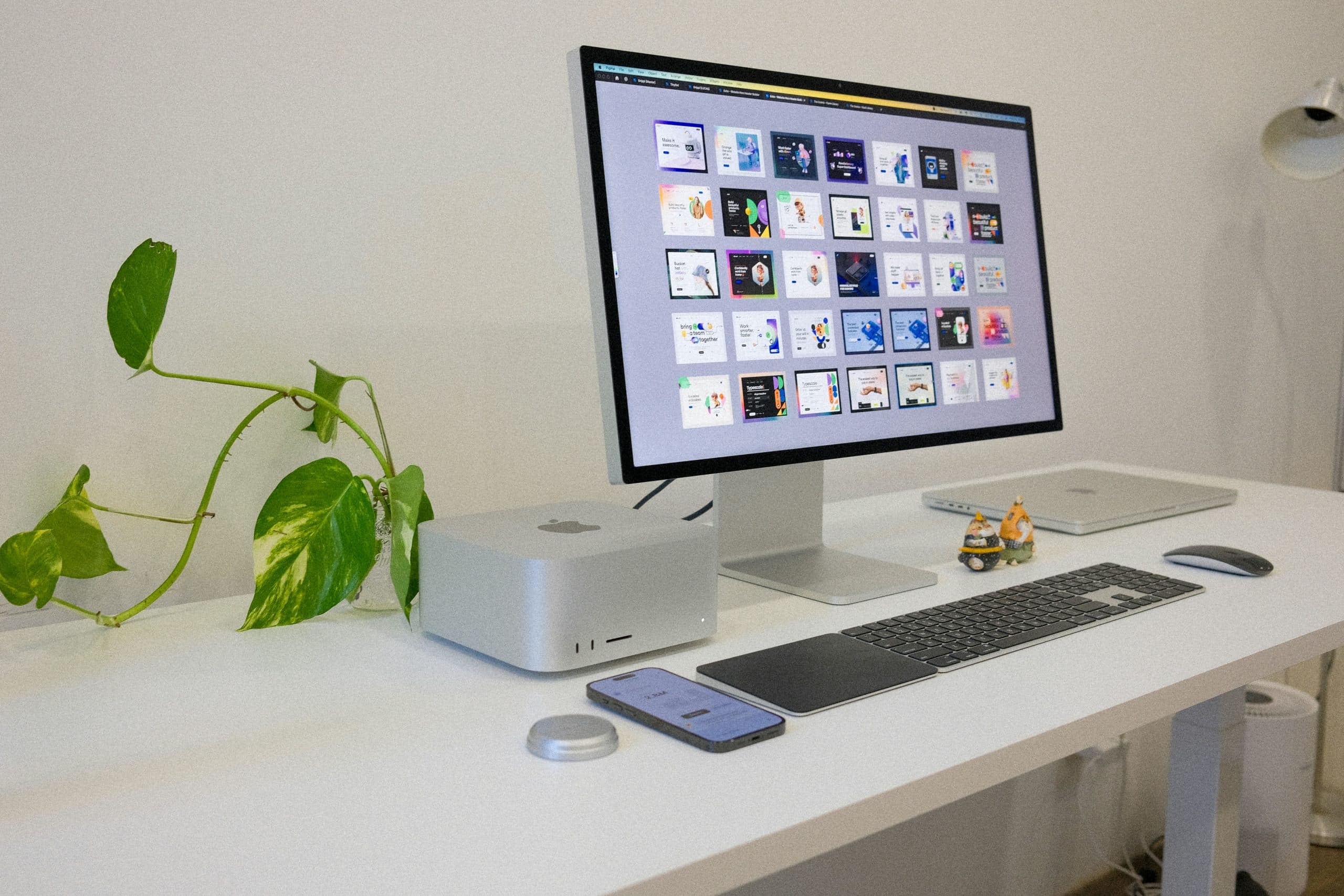 A modern workspace highlights a desktop monitor displaying a grid of thumbnail images, powered by a sleek M3 Ultra. Accompanying it on the white desk are a smartphone, keyboard, and mouse. A green potted plant and small figurines add charm beside the monitor.