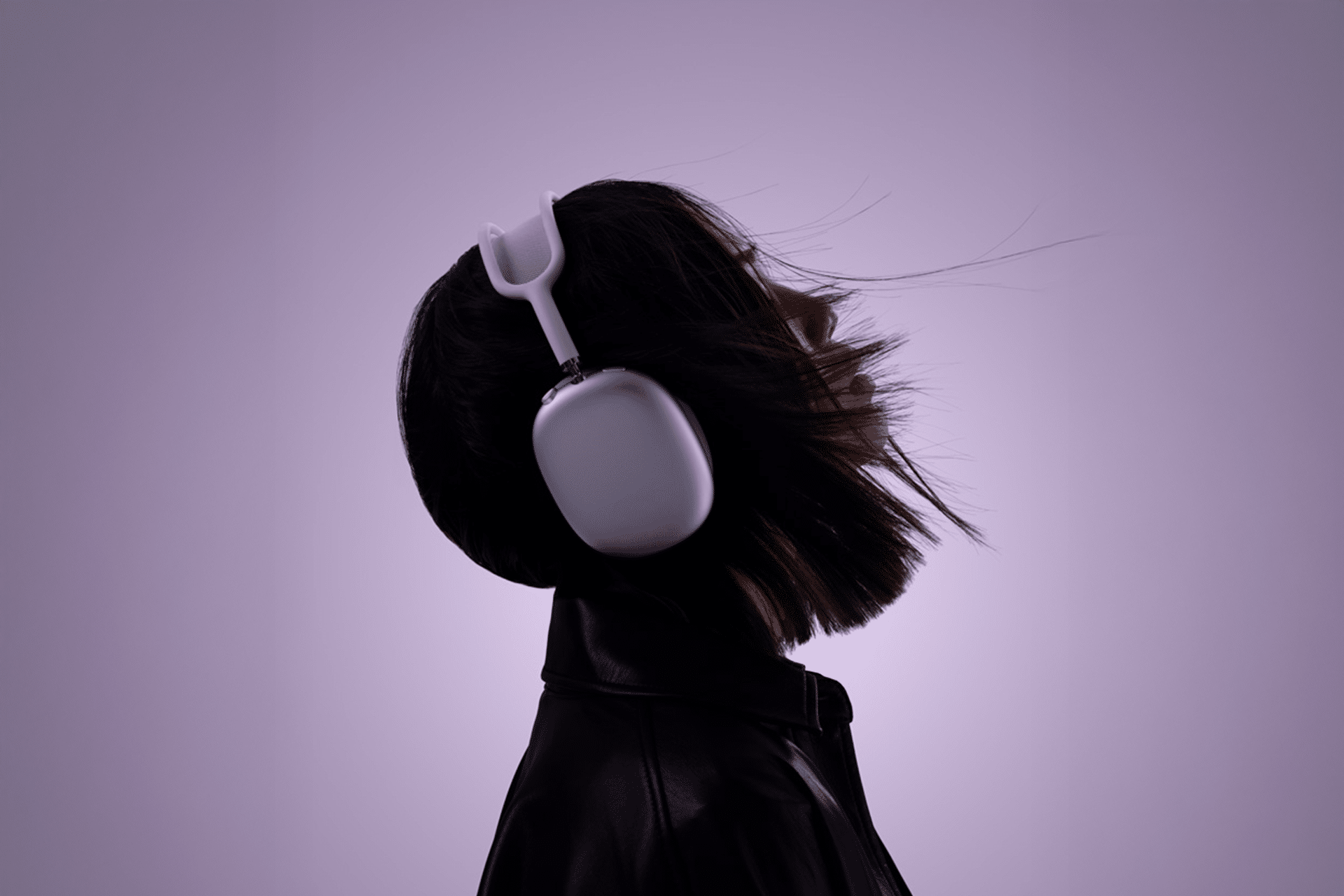 Side profile of a person wearing large over-ear AirPods Max, head tilted back, with short hair slightly flowing. The background is a soft purple gradient. The person is dressed in a dark jacket, creating a stylish, modern vibe.