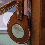 A brown leather keychain holder with white stitching is attached to a matching bag, securely holding an AirTag 2 for reliable tracking.