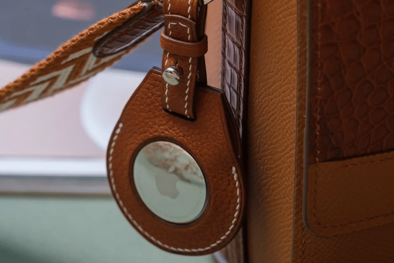 A brown leather keychain holder with white stitching is attached to a matching bag, securely holding an AirTag 2 for reliable tracking.