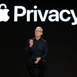 A person in dark clothing sits on a stool, gesturing while speaking on stage in front of a large screen displaying the Apple logo and the word "Privacy," highlighting iPhone security features.