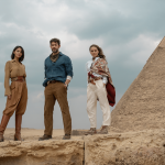 Three people stand on rocky terrain with pyramids in the background, embodying a timeless elegance as if inspired by the Fountain of Youth. They wear stylish, casual clothing; two women in boots and a man with a scarf. The cloudy sky adds drama as they look confidently towards the camera.