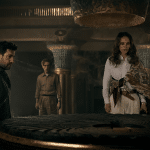 Three people stand around a large, circular stone artifact in a dimly lit, ancient-looking room. The woman in white examines the artifact closely, rumored to hold secrets of the Fountain of Youth, while the two men watch intently. Ornate columns and subdued lighting add to the mystique.