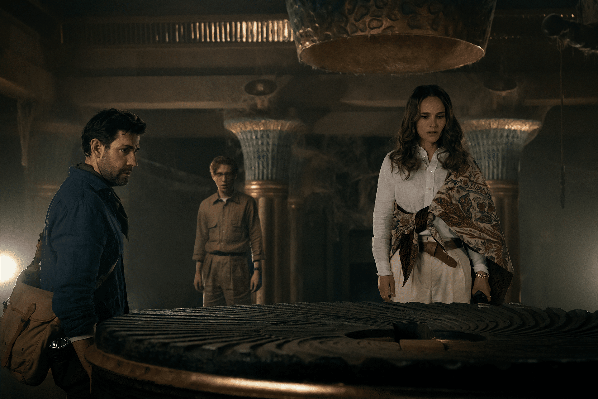 Three people stand around a large, circular stone artifact in a dimly lit, ancient-looking room. The woman in white examines the artifact closely, rumored to hold secrets of the Fountain of Youth, while the two men watch intently. Ornate columns and subdued lighting add to the mystique.