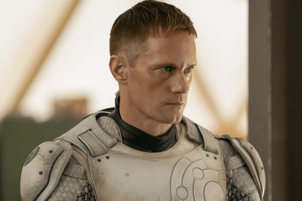 A person with short hair, clad in futuristic armor reminiscent of an Apple TV aesthetic, looks to the side. The light-colored armor features high-tech designs over a dark undershirt. Blurred in the background is an indoor setting with a metallic, industrial feel.