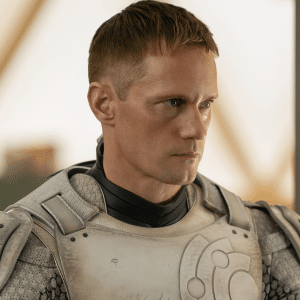 A person with short hair, clad in futuristic armor reminiscent of an Apple TV aesthetic, looks to the side. The light-colored armor features high-tech designs over a dark undershirt. Blurred in the background is an indoor setting with a metallic, industrial feel.