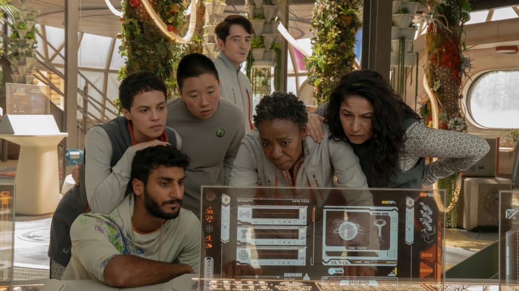 A diverse group of people wearing casual clothes huddles around a futuristic, illuminated screen displaying various data on Murderbot. The setting has greenery and modern technology, suggesting a high-tech environment. They appear focused and engaged.