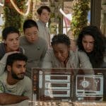 A diverse group of people wearing casual clothes huddles around a futuristic, illuminated screen displaying various data on Murderbot. The setting has greenery and modern technology, suggesting a high-tech environment. They appear focused and engaged.
