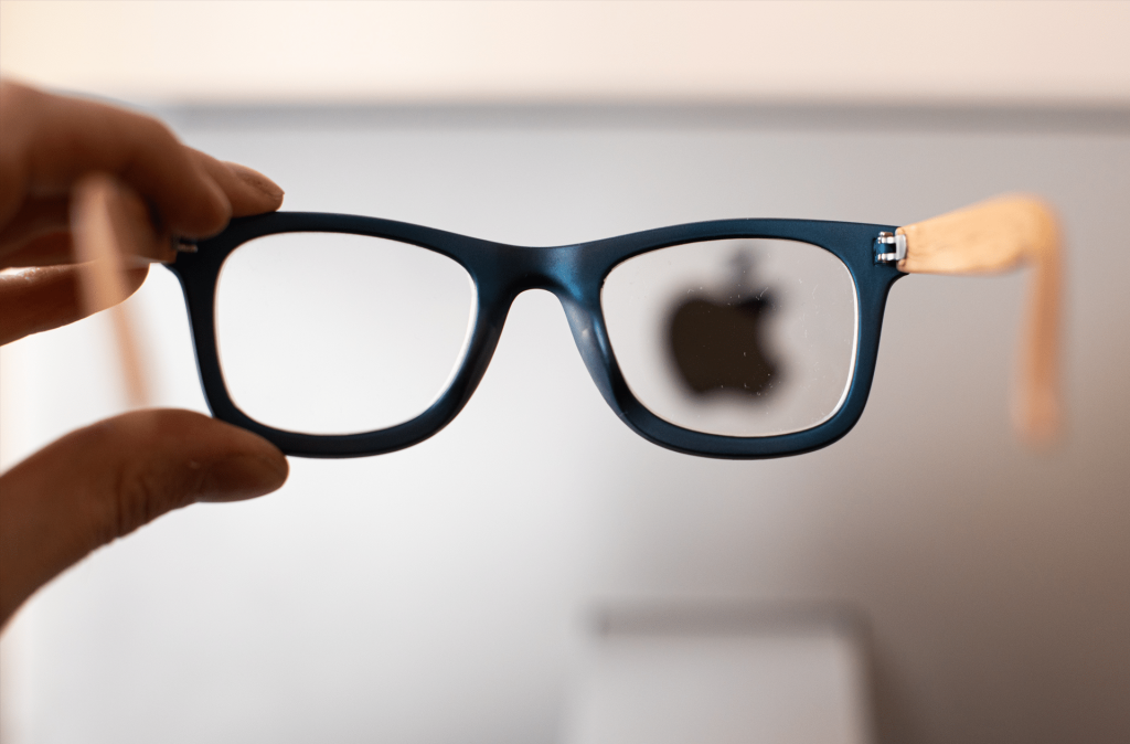 A hand holds up a pair of eyeglasses featuring N50 lenses in front of a blurred background, bringing the Apple logo into sharp focus through the right lens.