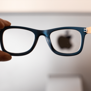 A hand holds up a pair of eyeglasses featuring N50 lenses in front of a blurred background, bringing the Apple logo into sharp focus through the right lens.