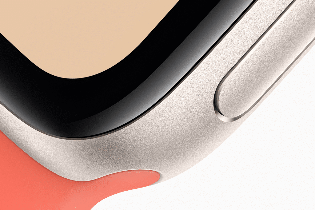 Close-up of the corner of an Apple Watch SE 3 with a metallic casing, black screen edge, side button, and a coral-colored strap.