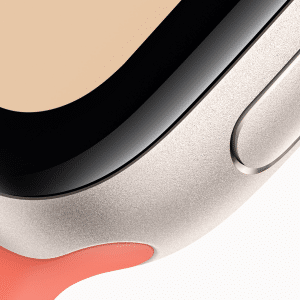 Close-up of the corner of an Apple Watch SE 3 with a metallic casing, black screen edge, side button, and a coral-colored strap.