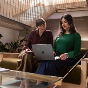 Two women sit on a couch in a modern office, looking at a laptop together. One woman holds a white cane, indicating she is visually impaired. There are plants and stairs in the background.
