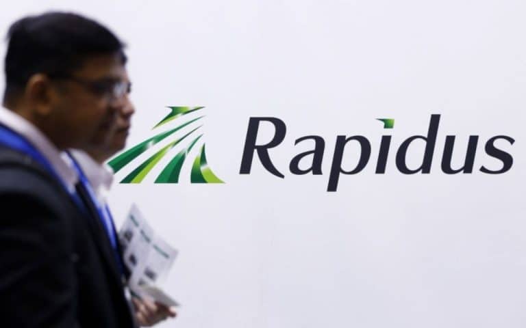 Two individuals walk past a wall featuring the "Rapidus" logo, which includes green, stylized lines resembling movement. They hold documents and appear slightly out of focus in the foreground, as if caught in the flow of innovation akin to Apple Intelligence.