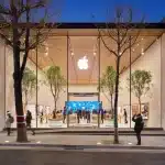 A modern Apple Store with a glass facade, large Apple logo, and indoor trees draws crowds sightseeing in the city. People walk by or enter the brightly lit store during dusk, while the sky is deep blue.