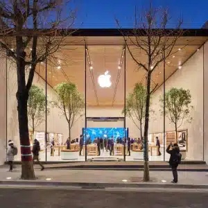 A modern Apple Store with a glass facade, large Apple logo, and indoor trees draws crowds sightseeing in the city. People walk by or enter the brightly lit store during dusk, while the sky is deep blue.