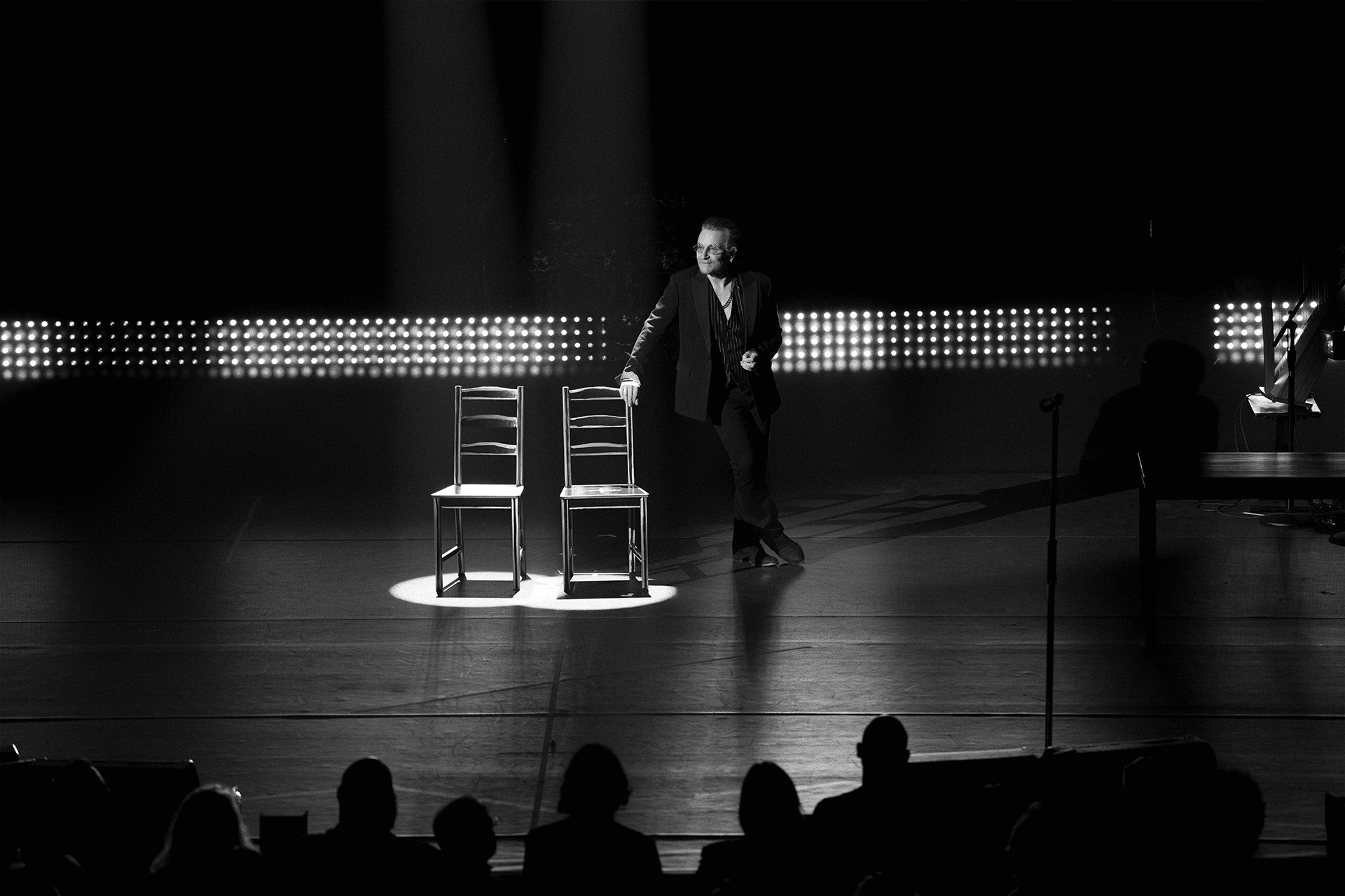 A man in a suit stands on a dimly lit stage beside two empty chairs, illuminated by a spotlight. Silhouetted audience members watch as bright lights form a backdrop—an evocative scene from an Apple TV+ summer 2025 release, in striking black and white.