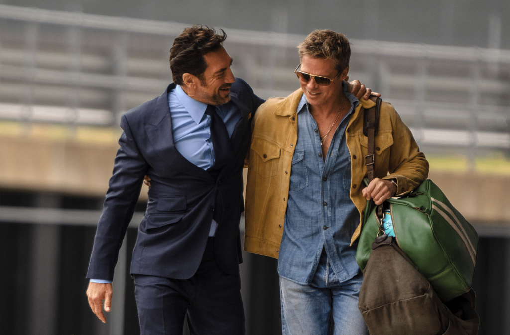 Two men walk together smiling; one in a blue suit with his arm around the other, who wears sunglasses, a tan jacket over a denim shirt, and carries two large bags—enjoying a friendly conversation outdoors in this Apple TV+ summer 2025 scene.