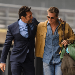 Two men walk together smiling; one in a blue suit with his arm around the other, who wears sunglasses, a tan jacket over a denim shirt, and carries two large bags—enjoying a friendly conversation outdoors in this Apple TV+ summer 2025 scene.