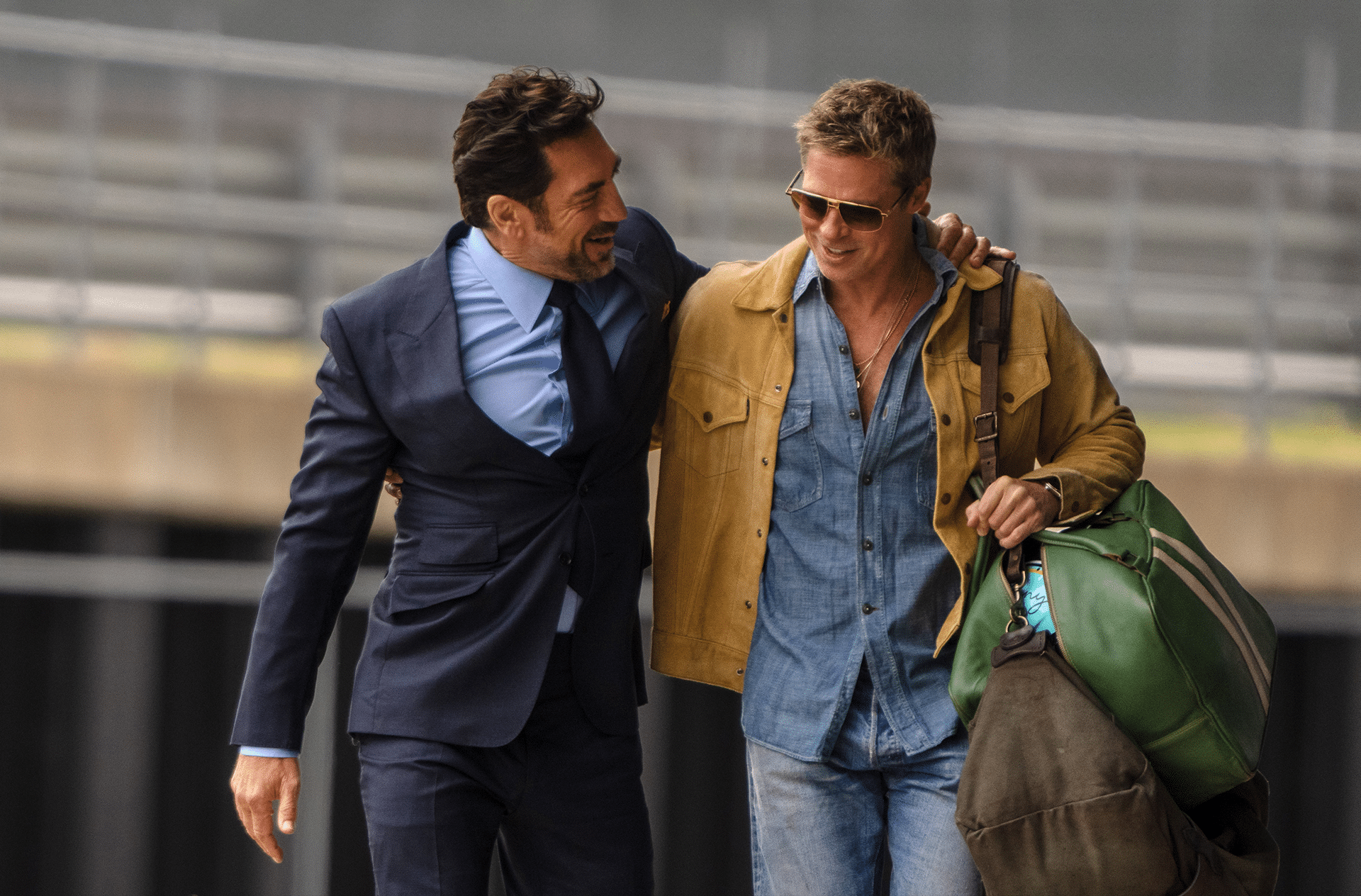 Two men walk together smiling; one in a blue suit with his arm around the other, who wears sunglasses, a tan jacket over a denim shirt, and carries two large bags—enjoying a friendly conversation outdoors in this Apple TV+ summer 2025 scene.