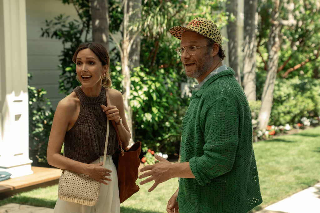 A woman in a sleeveless brown top and a man in a green sweater and leopard-print cap stand outside in a lush garden, both appearing engaged and mid-conversation, much like a scene from Platonic Season 2.