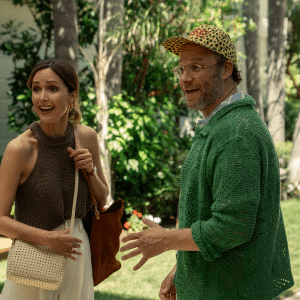 A woman in a sleeveless brown top and a man in a green sweater and leopard-print cap stand outside in a lush garden, both appearing engaged and mid-conversation, much like a scene from Platonic Season 2.