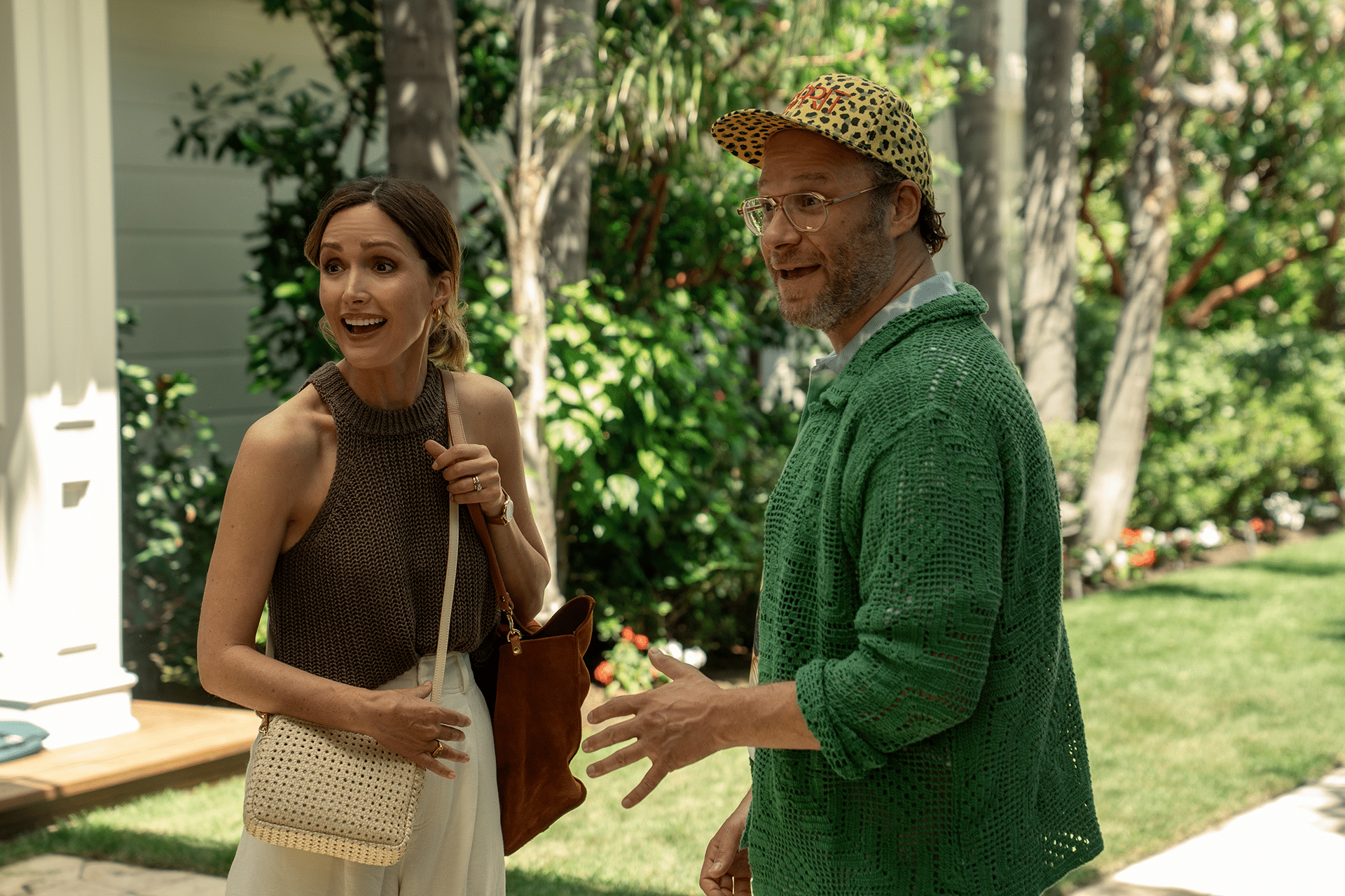 A woman in a sleeveless brown top and a man in a green sweater and leopard-print cap stand outside in a lush garden, both appearing engaged and mid-conversation, much like a scene from Platonic Season 2.