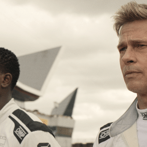 Two race car drivers in white suits stand side by side outdoors, looking focused. The background shows a racetrack and modern, angular buildings under a cloudy sky, evoking the excitement of an Apple TV+ motorsports series.