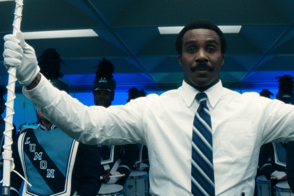 A man in a white shirt and striped tie stands with arms raised, holding a staff, in front of a Severance-inspired marching band dressed in blue uniforms with "LUMON" visible on one jacket. The background glows with blue light.