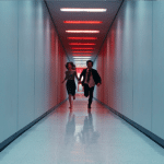 A man and woman, evoking a Severance-like atmosphere, run down a long, narrow hallway with white walls and a shiny floor, illuminated by red ceiling lights.