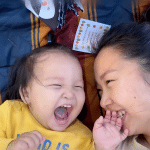 A baby and an adult woman lie on a striped blanket, laughing joyfully together. Captured as an Apple Vision Pro spatial video, their big smiles and playful moment are brought to life, with a card and toy nearby on the blanket.