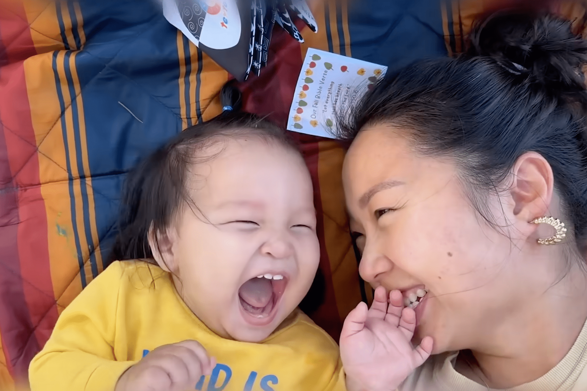 A baby and an adult woman lie on a striped blanket, laughing joyfully together. Captured as an Apple Vision Pro spatial video, their big smiles and playful moment are brought to life, with a card and toy nearby on the blanket.