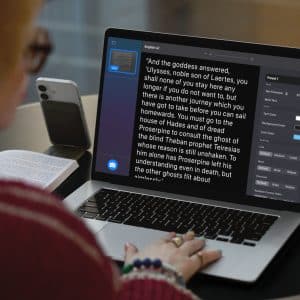 A person sits at a table using a laptop with a text-editing app open, displaying a passage and style options. An open book and a smartphone rest nearby on the table. The person's hands are visible on the laptop keyboard.